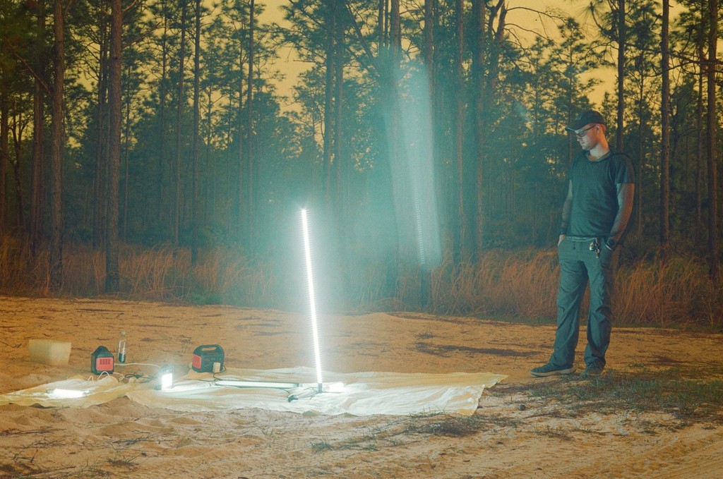 Nolan Webb standing beside a tall light on a stand over a white sheet in a sandy clearing, with portable power stations on the sheet and pine woods behind him